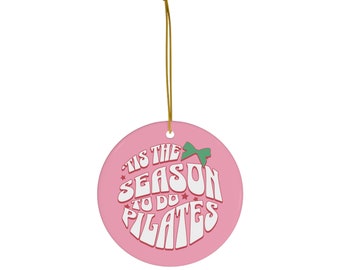 Könnte beinhalten: Rosafarbener Weihnachtsbaumschmuck aus Keramik mit einem weißen Design, auf dem "Tis the season to do Pilates" steht, mit einer grünen Schleife.