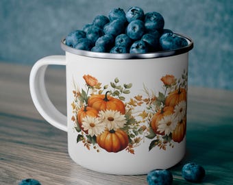 Puede incluir: Taza de esmalte blanco con un diseño de otoño con calabazas naranjas y flores blancas. La taza está llena de arándanos.