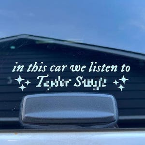 May include: White vinyl car decal with the text "in this car we listen to Taylor Swift" in a script font. The decal includes starburst graphics on either side of the text. The decal is affixed to a car window.