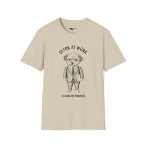 Puede incluir: Una camiseta beige con una ilustración en blanco y negro de un perro con traje y corbata. El texto "Clerk at Work" y "I know my folders" está impreso en la camiseta.