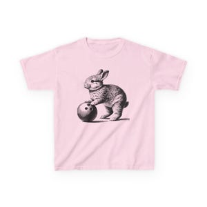 以下が含まれることがあります： ピンク色のTシャツ。白黒のイラストで、ウサギがボウリングの球を持っています。ウサギは陰影で詳細に描かれ、ボウリングの球には3つの穴があります。Tシャツは無地です。