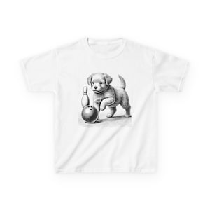 May include: White t-shirt featuring a black and white illustration of a puppy playing with a bowling ball and pin. The puppy is standing next to the bowling ball, with the pin in the background. The design is centered on the shirt.
