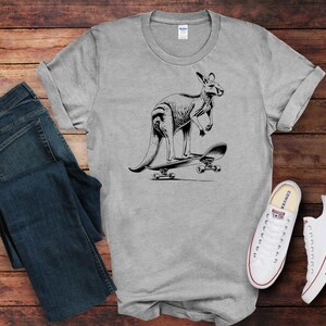 May include: A gray t-shirt with a black and white illustration of a kangaroo skateboarding. The kangaroo is standing on a skateboard with wheels and is holding onto the board with its front paws.
