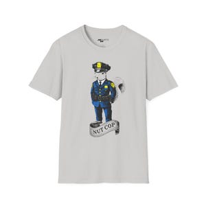 May include: A light gray t-shirt with a graphic of a squirrel dressed as a police officer. The squirrel is wearing a blue uniform with a yellow badge and a black belt. The text "NUT COP" is printed on a banner below the squirrel.