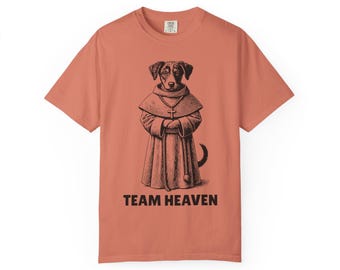 May include: Coral-colored t-shirt featuring a black and white illustration of a dog dressed as a monk, wearing a robe and a cross necklace. The text "TEAM HEAVEN" is printed in black below the illustration. The shirt has short sleeves and a crew neck.