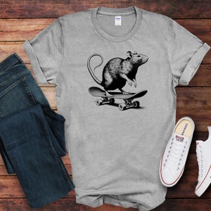 May include: A gray t-shirt with a black and white illustration of a rat skateboarding. The rat is standing on a skateboard with wheels and is looking to the right. The t-shirt is on a wooden surface with a pair of blue jeans and white sneakers.