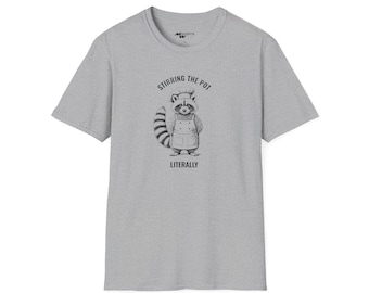 Op de afbeelding: Een grijs T-shirt met een afbeelding van een wasbeer die een koksmuts en een schort draagt. De tekst "Stirring the Pot Literally" staat onder de wasbeer.