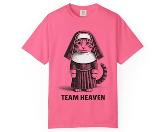 May include: A pink t-shirt featuring a graphic of a cat dressed as a nun, with the words "TEAM HEAVEN" printed below. The cat is depicted in a detailed, vintage style, wearing a habit and a cross. The shirt is a solid, vibrant pink color.