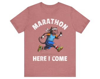 May include: A dusty rose t-shirt featuring a cartoon cat running a marathon. The cat wears a headband, wristbands, and running shoes. The words "MARATHON" and "HERE I COME" are printed above and below the cat illustration.