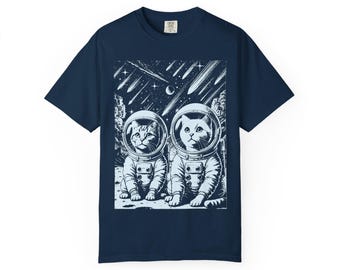 May include: Navy blue t-shirt featuring a black and white graphic of two cats in astronaut suits, set against a backdrop of a starry sky and shooting stars. The cats are sitting on a planet's surface.