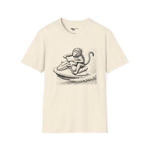 May include: A beige t-shirt with a black and white illustration of a monkey riding a jet ski. The monkey is wearing a life jacket and holding onto the handlebars. The jet ski is leaving a wake behind it.
