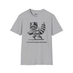 Puede incluir: Una camiseta gris con una ilustración en blanco y negro de un mapache con un casco de fútbol y sosteniendo un balón de fútbol. El texto "It's all about blocking and tackling" está impreso debajo de la ilustración.