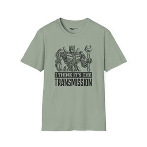 May include: A light green t-shirt with a black and white illustration of a robot holding a wrench. The text "I think it's the transmission" is printed below the robot.