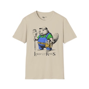 Puede incluir: Una camiseta beige con un castor de dibujos animados que lleva un mono y sostiene una llave inglesa. El castor lleva una camisa verde y tiene un cinturón de herramientas con herramientas. El texto "Lord of the Wax Rings" está impreso debajo del castor.