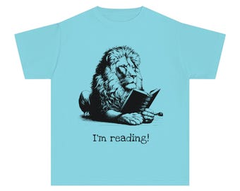 May include: Light blue t-shirt featuring a black and white illustration of a lion reading a book. The text "I'm reading!" is printed below the lion. The shirt is made of a soft material.