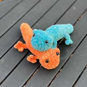 May include: Two crocheted plush lizards, one blue with white speckles and one orange with white speckles, are stacked on top of each other on a black wooden surface.