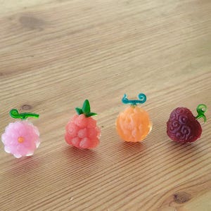 Peut inclure: Quatre figurines translucides en forme de fruits, roses, corail, orange et rouge foncé. Chacune a une tige verte ou turquoise. Elles sont disposées sur une surface en bois brun clair.