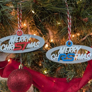 May include: Two Christmas tree ornaments with the words "MERRY CHRISTMAS" in white and red or blue lettering. The ornaments are gray ovals with a red and white string for hanging. The ornaments are hanging on a Christmas tree.