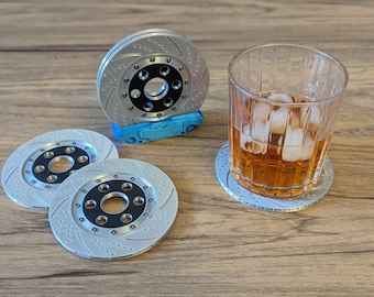 Puede incluir: Un juego de cuatro posavasos plateados con forma de rotor de freno de coche y centros negros. Un posavasos sostiene un vaso de líquido ámbar con hielo. Los posavasos están sobre una superficie de madera.