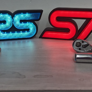 May include: Two illuminated car emblems, one blue and one red, spelling out "RS" and "ST" respectively. The emblems are on a light wood surface with two wrenches and a socket.