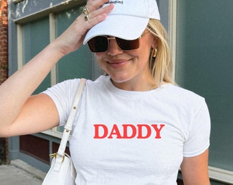 May include: A white t-shirt with the word "DADDY" printed in red letters. The person is wearing blue jeans and a white baseball cap.
