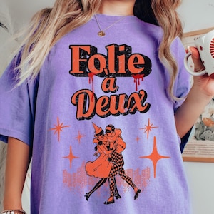 May include: Purple t-shirt with a vintage-style graphic of two people dancing. The graphic says "Folie a Deux" in orange and red lettering.