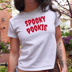 Może przedstawiać: Jasnoszara koszulka z czerwonym napisem "SPOOKY POOKIE". Koszulka ma okrągły dekolt i wydaje się być wykonana z miękkiego materiału. Słowo "ASH" jest wydrukowane w prawym dolnym rogu.