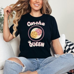 May include: A black t-shirt with a graphic of two pink and yellow conchas, a traditional Mexican sweet bread, with the text "Concha Queen" in a retro font.