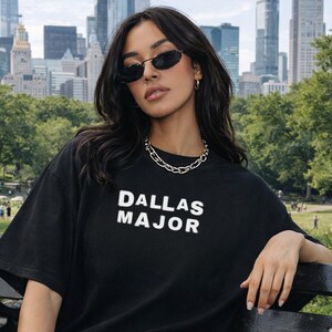 May include: Black t-shirt with the text "DALLAS MAJOR" in white block letters. The model is wearing sunglasses and a silver chain necklace. The word "BLACK" is at the bottom right corner of the image.