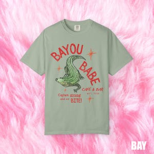 Op de afbeelding: Saliegroen t-shirt met een afbeelding van een groene alligator en de tekst "BAYOU BABE CAFE & BAR EST. 1996". De tekst "Cajun strong and we BITE!" staat er ook op. De achtergrond is roze en pluizig.