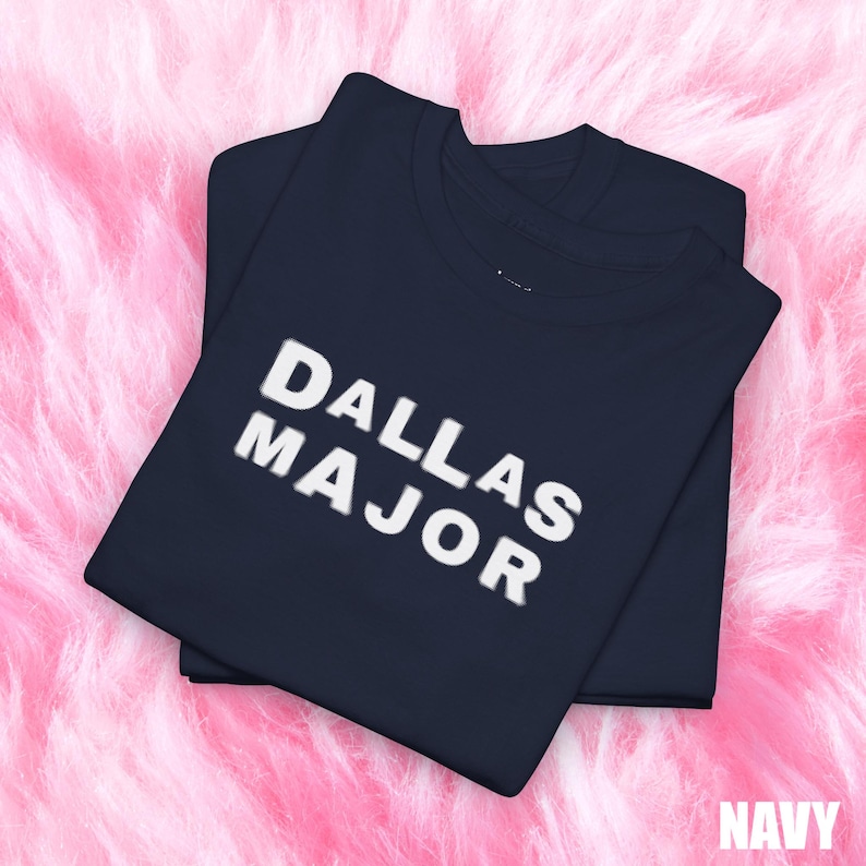May include: Two navy blue t-shirts are stacked on a pink, fluffy surface. The top shirt displays the text "DALLAS MAJOR" in white, bold letters. The word "NAVY" is visible in the bottom right corner.