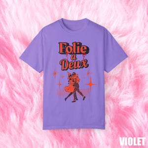 May include: A purple t-shirt with a graphic print of two people dancing. The text "Folie a Deux" is printed in red and black above the graphic.