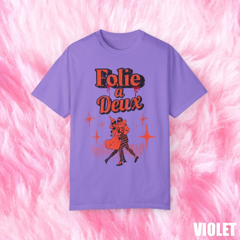 May include: A purple t-shirt with a graphic print of two people dancing. The text "Folie a Deux" is printed in red and black above the graphic.