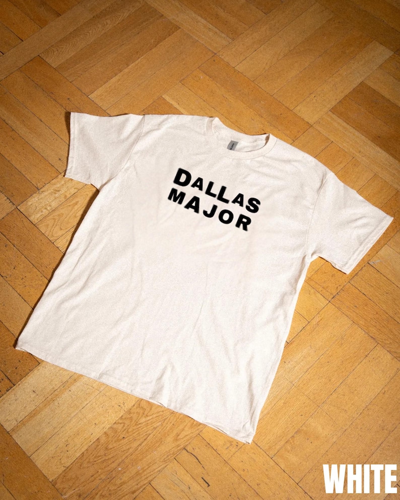 May include: A white t-shirt with the words "DALLAS MAJOR" printed in black, centered on the front. The shirt is laid flat on a wooden floor. The word "WHITE" is visible in the bottom right corner.