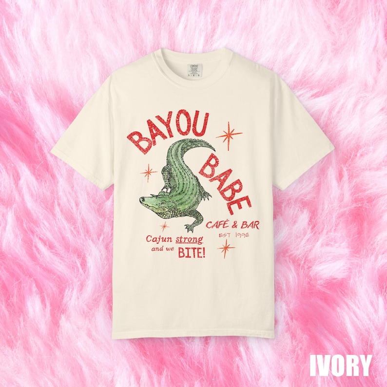Op de afbeelding: Ivoor t-shirt met een groen alligatorontwerp en de rode woorden "BAYOU BABE". Het shirt heeft ook de tekst "Cajun strong and we BITE!" en "CAFE & BAR EST 1996".