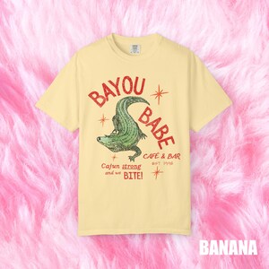 Op de afbeelding: Een lichtgeel t-shirt met de tekst "BAYOU BABE" in het rood, met een groene alligator illustratie. Het shirt bevat ook de tekst "Cajun strong and we BITE!" en "CAFE & BAR EST 1996". Het woord "BANANA" staat onderaan.