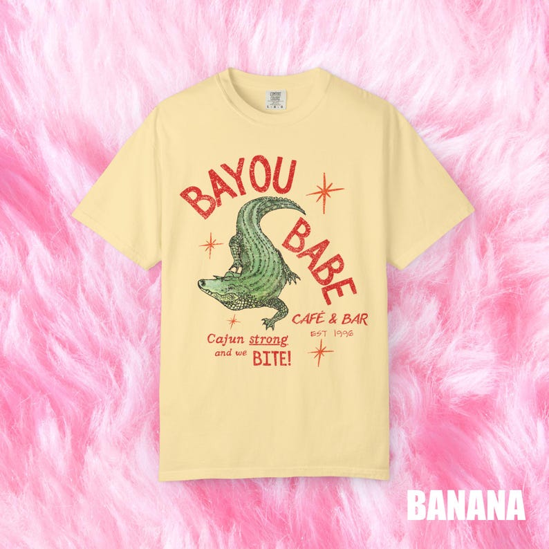 Op de afbeelding: Een lichtgeel t-shirt met de tekst "BAYOU BABE" in het rood, met een groene alligator illustratie. Het shirt bevat ook de tekst "Cajun strong and we BITE!" en "CAFE & BAR EST 1996". Het woord "BANANA" staat onderaan.
