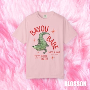 Op de afbeelding: Lichtroze t-shirt met rode tekst "BAYOU BABE" boven een groene alligator illustratie. De tekst "Cajun strong and we BITE!" en "CAFE & BAR EST 1996" staan er ook op.