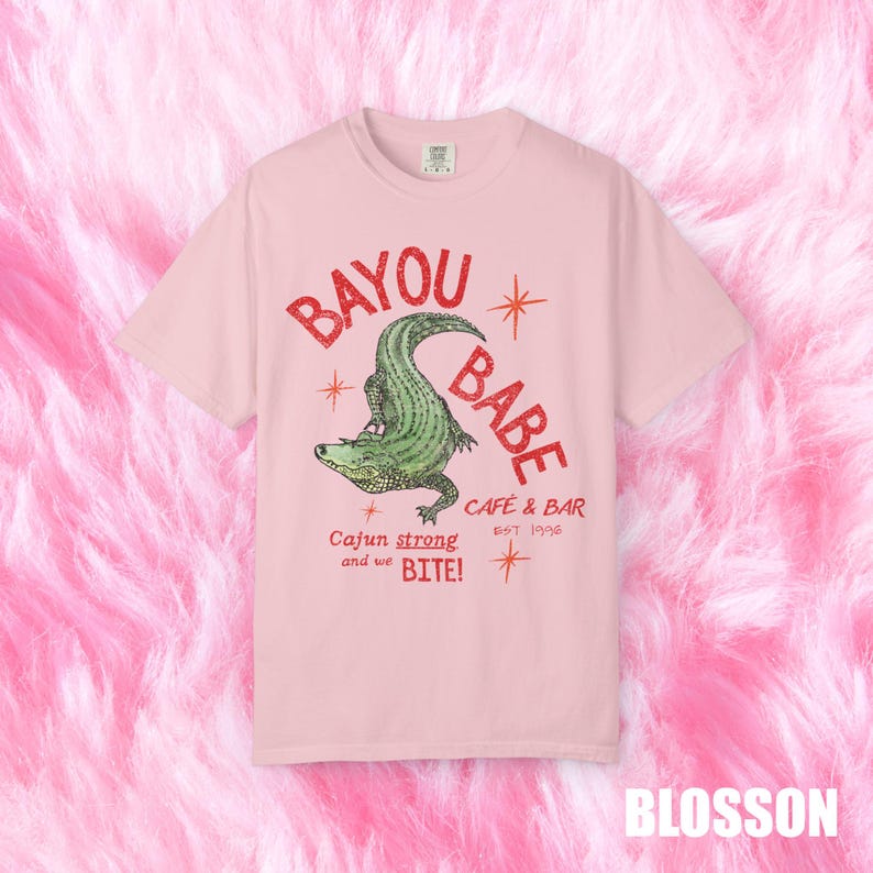 Op de afbeelding: Lichtroze t-shirt met rode tekst "BAYOU BABE" boven een groene alligator illustratie. De tekst "Cajun strong and we BITE!" en "CAFE & BAR EST 1996" staan er ook op.