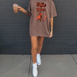 May include: A brown oversized t-shirt with a graphic print of a couple dancing. The graphic text reads "Folie a Deux" in orange with a red drip effect. The model is wearing white high-top sneakers.