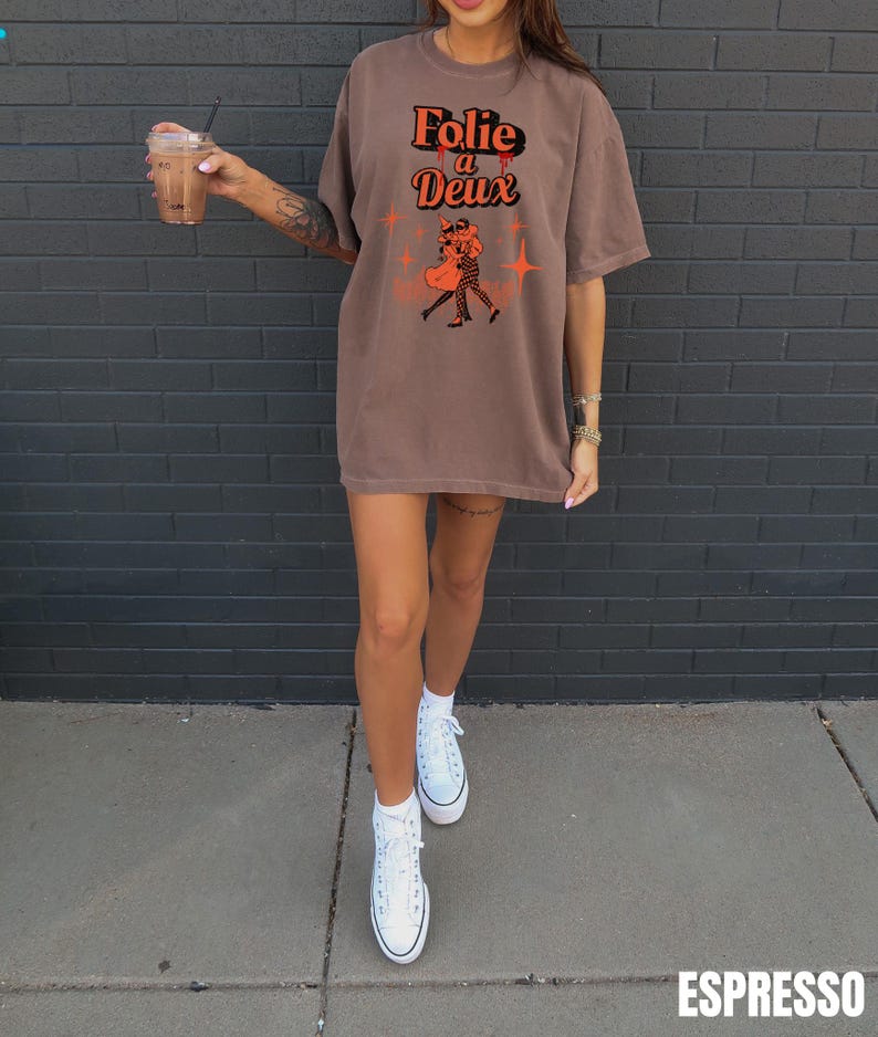 May include: A brown oversized t-shirt with a graphic print of a couple dancing. The graphic text reads "Folie a Deux" in orange with a red drip effect. The model is wearing white high-top sneakers.