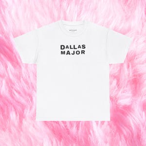 May include: White t-shirt with the text "DALLAS MAJOR" in black block letters. The shirt has a crew neck and short sleeves. The background is a soft, pink, fluffy texture.