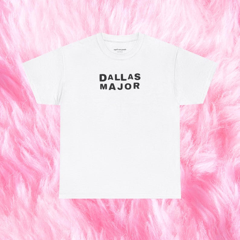 May include: White t-shirt with the text "DALLAS MAJOR" in black block letters. The shirt has a crew neck and short sleeves. The background is a soft, pink, fluffy texture.