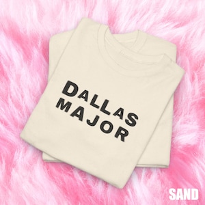 May include: Two sand-colored t-shirts are neatly folded and stacked. The shirts feature the text "DALLAS MAJOR" in bold, black letters. The word "SAND" is printed in the bottom right corner. The shirts are on a pink, fluffy surface.