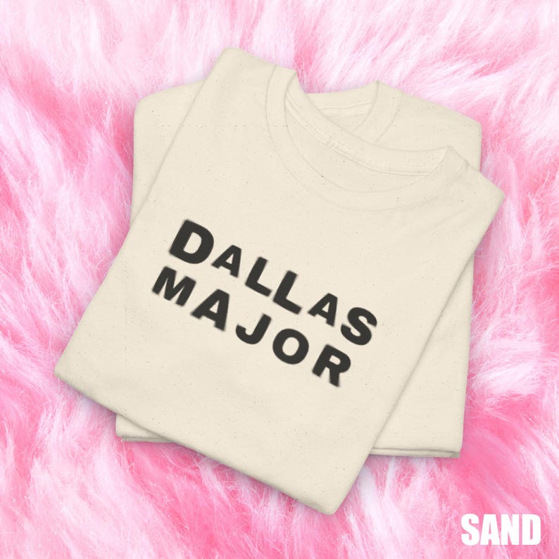 May include: Two sand-colored t-shirts are neatly folded and stacked. The shirts feature the text "DALLAS MAJOR" in bold, black letters. The word "SAND" is printed in the bottom right corner. The shirts are on a pink, fluffy surface.