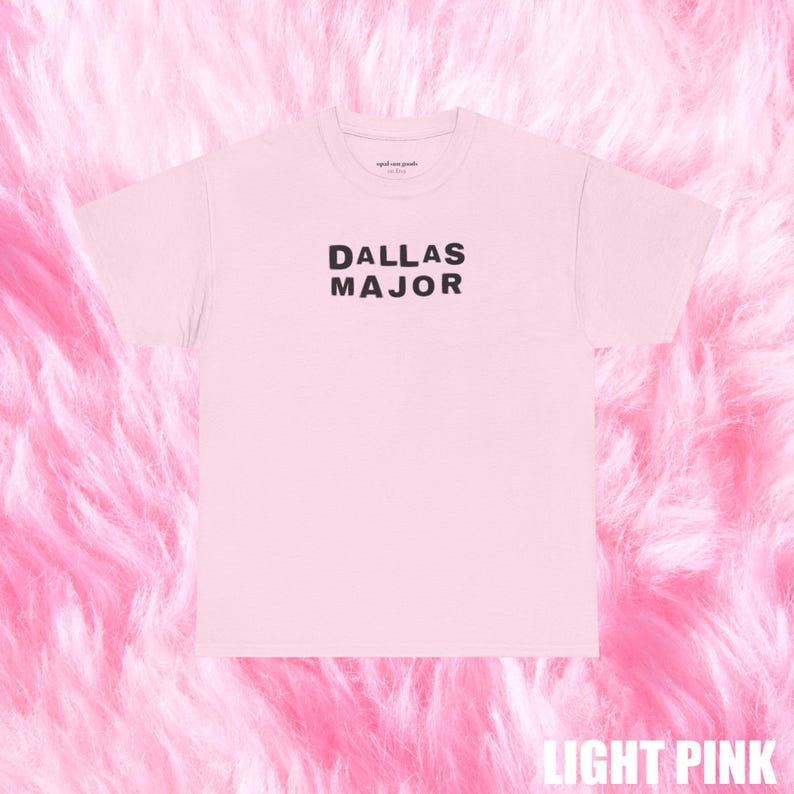 May include: A light pink t-shirt with the words "DALLAS MAJOR" printed in black. The shirt is displayed on a pink fluffy surface. The words "LIGHT PINK" are at the bottom of the image.
