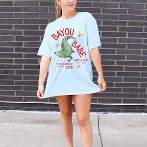 Op de afbeelding: Lichtblauw T-shirt met een afbeelding van een groene alligator en de tekst "Bayou Babe Cafe & Bar". Het shirt heeft ook de tekst "Cajun strong and we BITE!" en is gemaakt van zacht materiaal.