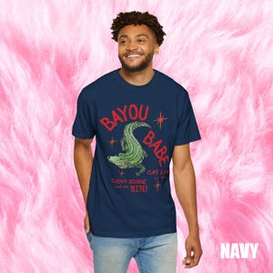 Op de afbeelding: Marineblauw T-shirt met de rode woorden "BAYOU BABE" en een groene alligatorafbeelding. Het shirt heeft ook de tekst "Cajun strong and we BITE!" en "CAFE & BAR EST. 2020".