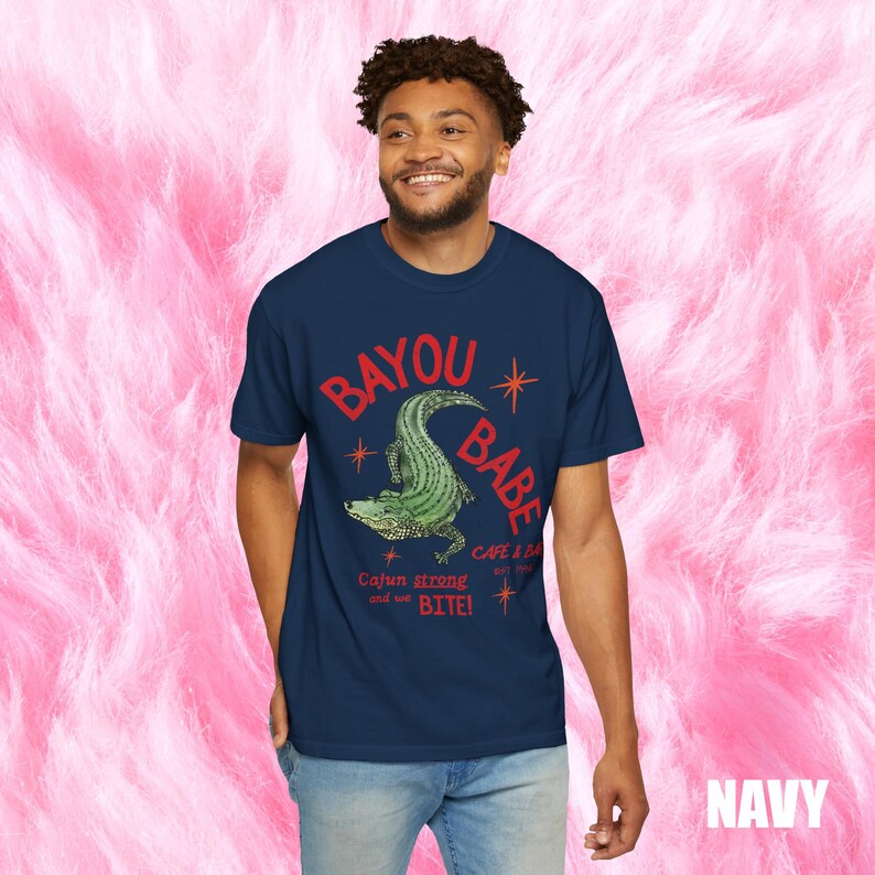 Op de afbeelding: Marineblauw T-shirt met de rode woorden "BAYOU BABE" en een groene alligatorafbeelding. Het shirt heeft ook de tekst "Cajun strong and we BITE!" en "CAFE & BAR EST. 2020".