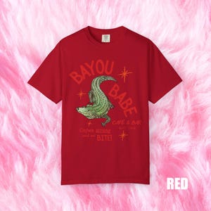 Op de afbeelding: Rood T-shirt met de tekst "BAYOU BABE" en een groene alligatorafbeelding. Het shirt bevat ook de tekst "Cajun strong and we BITE!" en "CAFE & BAR EST. 1996".